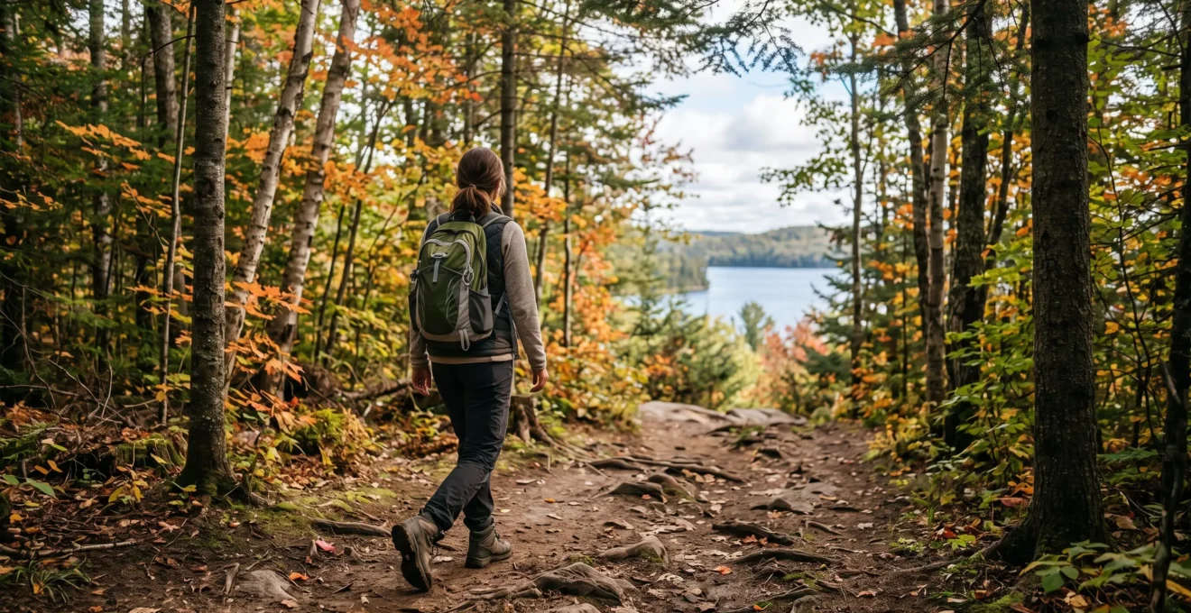 Une randonneuse marche sur un sentier forestier en automne dans un parc canadien, vêtue de couches de vêtements superposés.